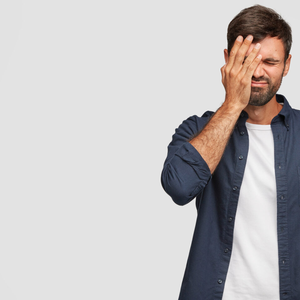 Indoor shot of handsome stressful overworked man covers face with palm, has displeased expression, dressed in casual clothes, poses against white background with copy space for your promotional text