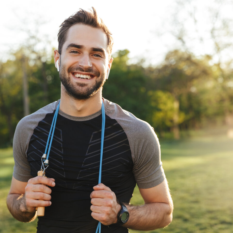 Image of a handsome young strong sports man posing outdoors at the nature park location with skipping rope.