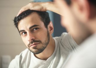 Latino person with beard grooming in bathroom at home. White metrosexual man worried for hair loss and looking at mirror his receding hairline.