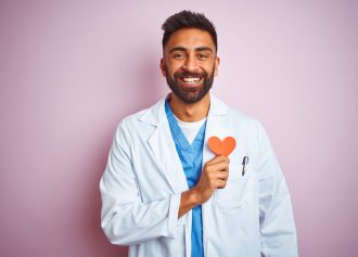 Young indian doctor man holding paper heart standing over isolated pink background with a happy face standing and smiling with a confident smile showing teeth